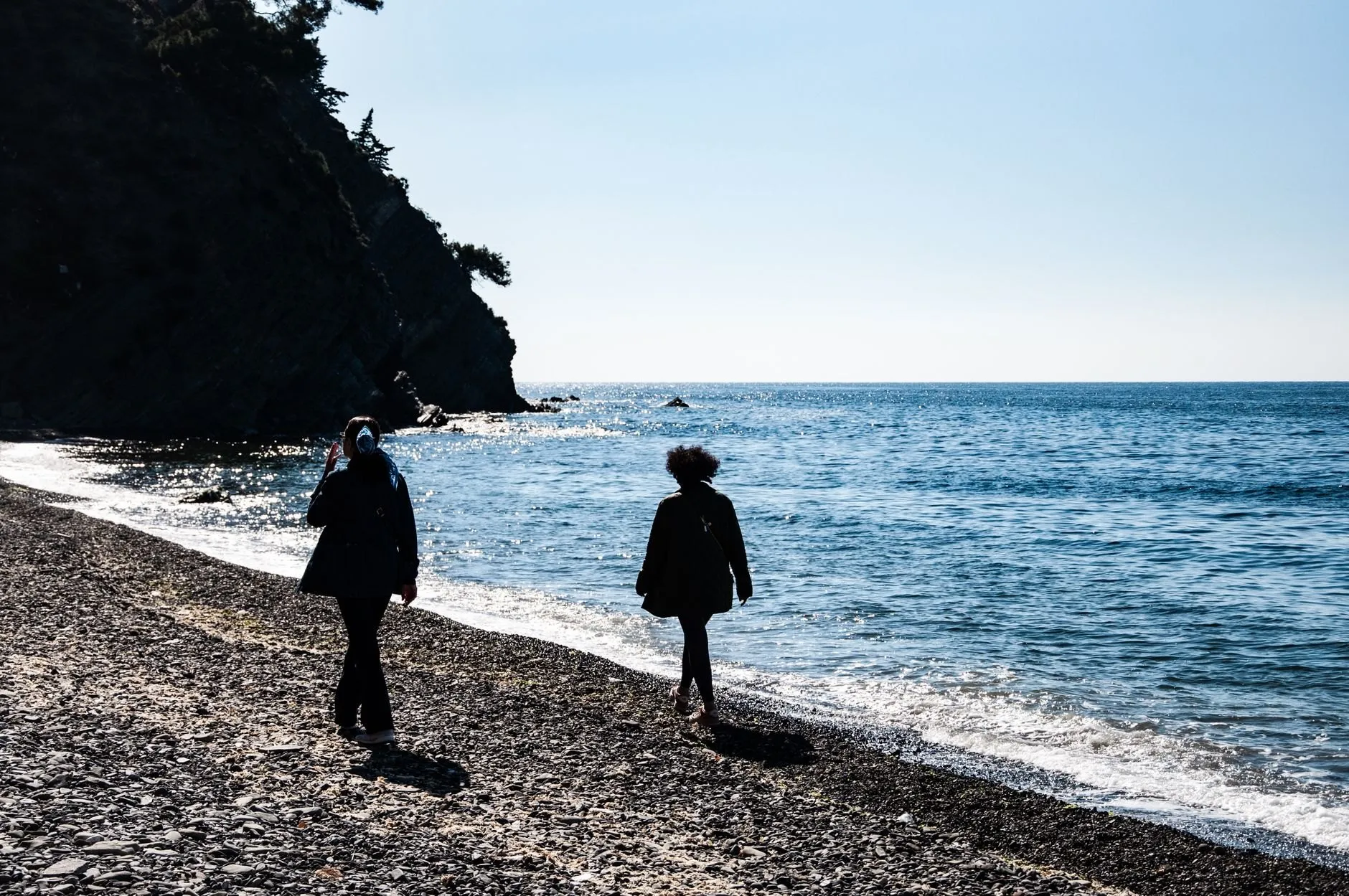 Two people walking along a shingle beach by a calm sea, seen from behind